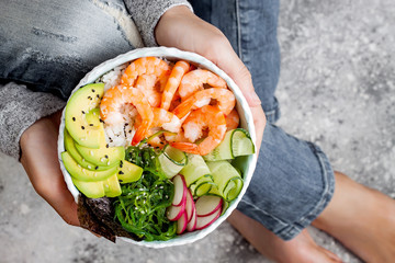 Girl in jeans holding shrimp poke bowl with seaweed, avocado, cucumber, radish, sesame seeds.
