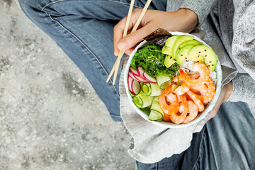 Girl in jeans holding shrimp poke bowl with seaweed, avocado, cucumber, radish, sesame seeds.