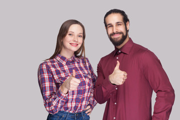 Portrait of happy satisfied bearded man with black collected hair and woman in casual style standing and looking at camera, smiling with thumbs up. indoor studio shot, isolated on gray background.