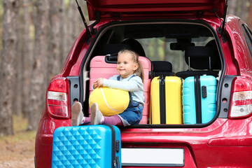 Cute little girl sitting in car trunk loaded with suitcases outdoors