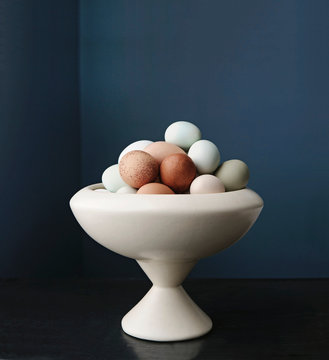 Fresh multicolored eggs are in a bowl in front of a dark blue wall.