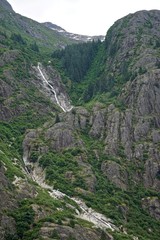 Endicott Arm, Alaska, USA: A waterfall flows down from a snow-capped mountain through a green valley in a fjord in the Pacific Northwest.