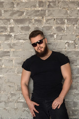 Fototapeta premium Bodybuilder in sunglasses portrait. Muscular man with a beard in the tight-fitting T-shirt standing at the brick wall. Stylish and sporty young man.