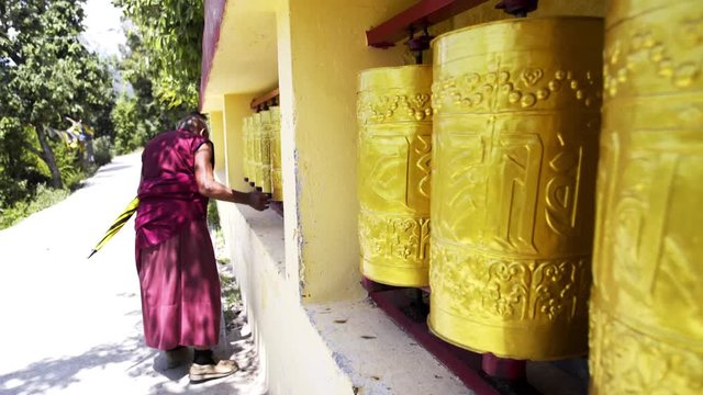 Back view unrecognizable senior buddhist monk touching and rotating colorful golden prayer wheels in residence of Dalai Lama in Dharamsala