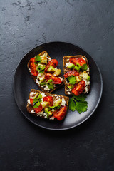 Avocados, cherry tomatoes and feta cheese sandwiches with balsamic sauce and cilantro on old olive board on dark black background. Top view.
