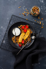 Oatmeal granola with yoghurt, fresh strawberries and banana, chia seeds, sunflower and honey in black ceramic plate on dark background. Top view.