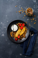 Oatmeal granola with yoghurt, fresh strawberries and banana, chia seeds, sunflower and honey in black ceramic plate on dark background. Top view.