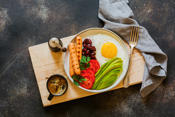 Traditional English breakfast with fried eggs, avocado, sausages grill, beans, and tomatoes on dark concrete background. Top view.