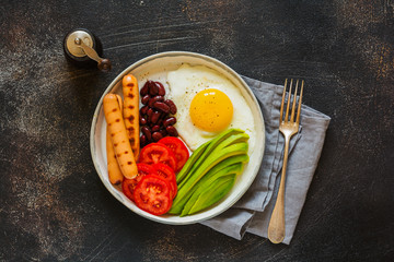 Traditional English breakfast with fried eggs, avocado, sausages grill, beans, and tomatoes on dark concrete background. Top view.