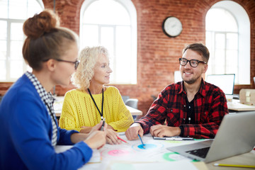 Group of creative people in casualwear sitting by table and discussing organization moments