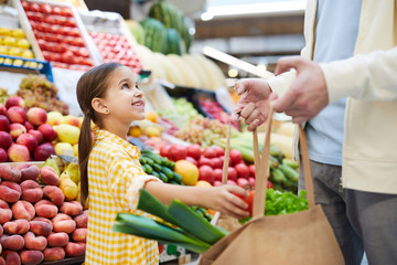 Cheerful girl in yellow dress putting vegetable into bag and looking at father while helping him with shopping at farmers market