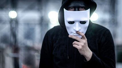 Mystery hoodie man with broken black mask holding white mask in his hand. Anonymous social masking...