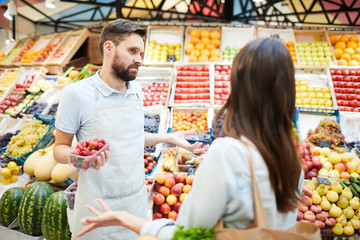 Confused misunderstanding young male grocer in apron holding container of strawberries shrugging shoulders while talking to customer in farmers market