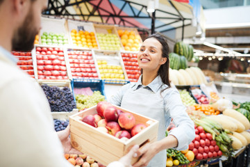 Cheerful content attractive young female grocer in apron giving box of apples to customer in farmers market, she working in organic food store