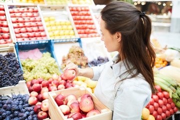 Serious pensive young female seller in apron placing products on food stall and choosing place for apples at farmers market