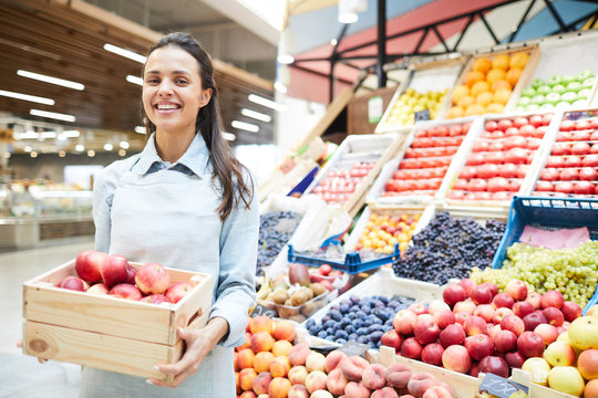Cheerful excited young female grocery retailer in apron standing against food stall and holding box of apples