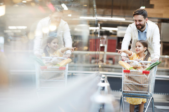 Positive girl pointing at product on shelf while pushing shopping cart together with handsome father in supermarket