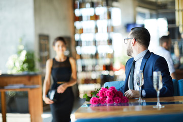 Man in suit sitting by table and looking at his girlfriend moving to him for date