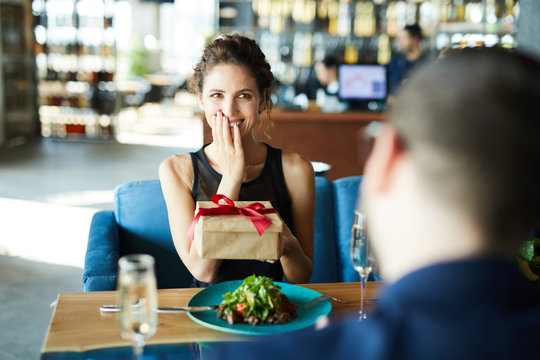 Young excited woman holding box with unexpected gift from her boyfriend during lunch in restaurant