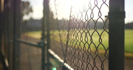 A chain link fence gate with locks on it at sunrise outside of a grass baseball field in a public park.