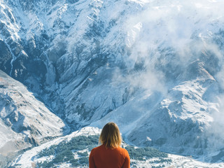 Traveler girl looking at the snowy mountains, standing back in a beautiful orange dress. Travel...