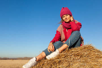 Naklejka na ściany i meble The concept of livestyle outdoor in autumn. Close up of a young woman student in a warm autumn clothes looking funny, smilling, posing for the camera, sitting on haystick