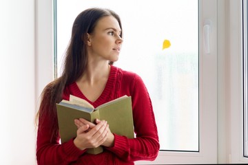 Young woman reading book near rainy autumn window with yellow leaf