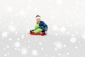 childhood, sledging and season concept - happy little boy sliding on sled down snow hill outdoors...