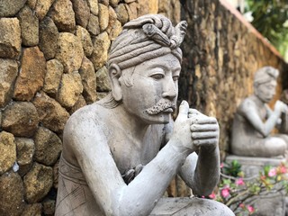 Stone statue of balinese priest with mustache praying while sitting crossed legs with pink flowers in the background on front of creme colored stone wall
