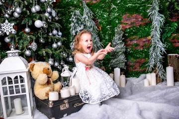 cute little girl in a white dress sitting near a Christmas tree on a suitcase next to the candles and a teddy bear, throws snow up and laughs