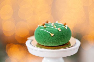 Mini mousse pastry dessert with green velour on garland lamps bokeh background. Modern european cake. French cuisine. Christmas theme