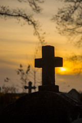 a cross on the background of the sunset and branches of the trees, a mysterious atmosphere. A weathered stone Christian cross silhouetted against an orange sunset.