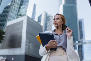 Beautiful young woman with folders and documents in her hands standing among the skyscrapers of the modern business center
