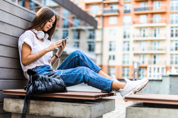 Young pretty woman sitting on a bench in the city. Young beautiful woman listening to music with phone in outdoors.
