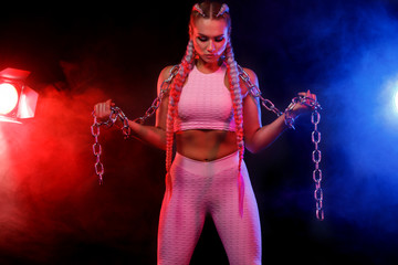 Sporty fit woman dancer and athlete with chains makes fitness exercising on black background with colorful lights.