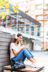 Fototapeta premium Portrait of a smiling girl with earphones listening to music sitting on the bench outdoors