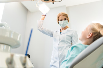 bottom view of young female dentist with lamp standing over little child in chair