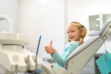 happy little child sitting in dental chair at dentist office and showing thumb up