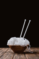 close up view of white yarn in bowl and knitting needles on wooden surface on black backdrop