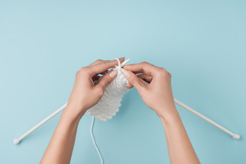 partial view of woman with white yarn and knitting needles knitting on blue backdrop