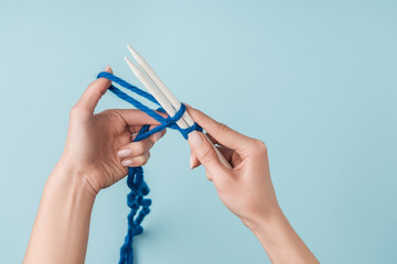 partial view of woman with blue yarn and white knitting needles knitting on blue backdrop