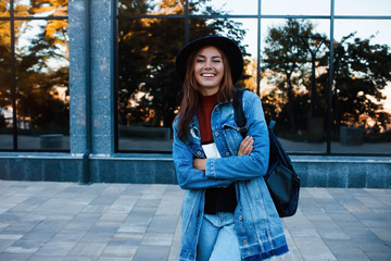 Hipster girl wearing casual sweater, jeans and backpack posing against glass street wall, minimalist urban clothing style.Stylish happy young woman wearing boyfriend jeans and long jeans coat.