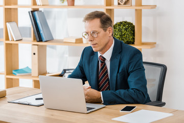 adult serious businessman in glasses sitting at table and using laptop in office