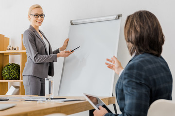 young smiling woman pointing at white board and man sitting at table with tablet in office