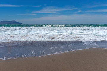Beautiful turquoise waves with foam on a sandy beach, against the blue sky with clouds and a mountain on the horizon