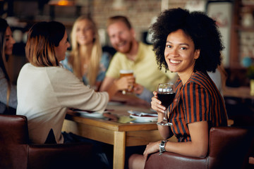 Woman looking at camera and holding glass of wine while sitting in restaurant. In background friends drinking and chatting.