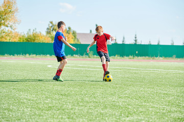 Obraz premium Full length portrait of two boys playing football in outdoor stadium lit by sunlight, copy space