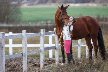 Young girl standing with her bay horse near paddock. Equestrian life concept background