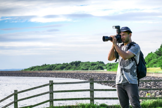 Closeup of Young handsome Professional photographer in white shirt with black backpack, taking landscape photos on the beach close to a wooden fence in a green nature with blue cloudy sky