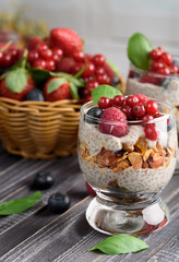 Two glasses of chia pudding with fresh strawberries, raspberries and blueberries. Basket with berries. On a wooden grey background.